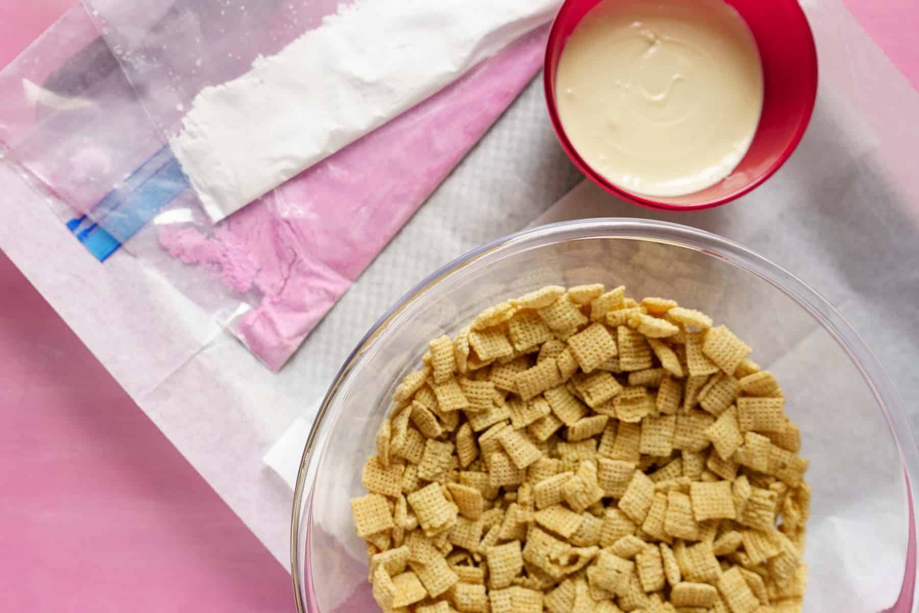 A bowl of cereal squares, a dish of melted white chocolate, and bags with powdered mixes are artfully arranged on a pink surface, creating the perfect setup for making Valentine puppy chow.