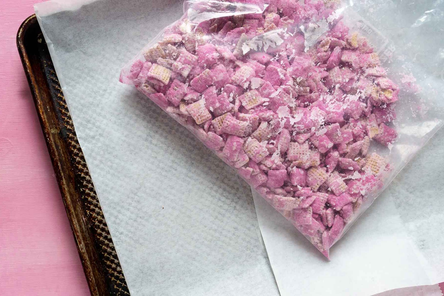 A delightful bag of pink-coated cereal squares, perfect for Valentine's Day, sits invitingly on a parchment-lined baking tray. This irresistible Valentine puppy chow is a treat that adds charm and sweetness to any occasion.