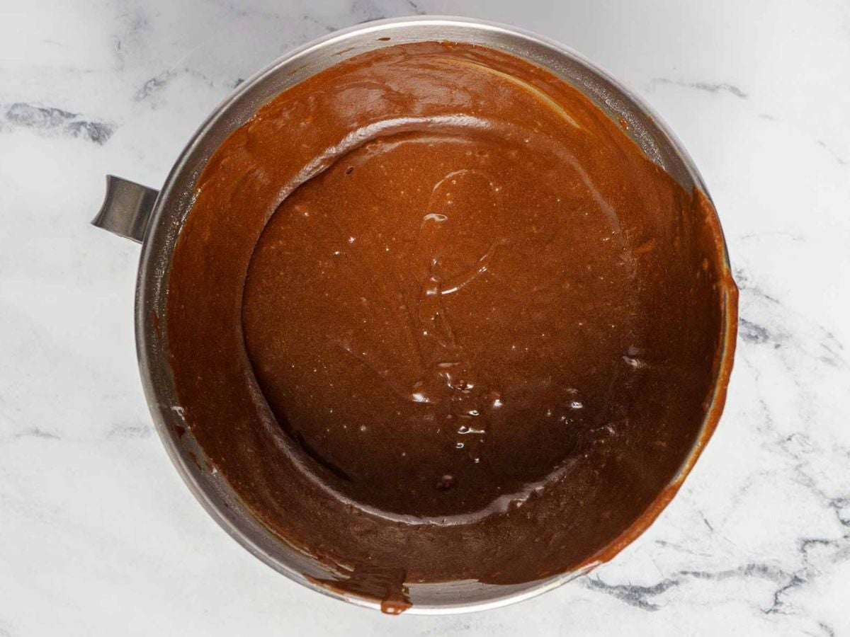 A metal mixing bowl filled with smooth, glossy gluten free dark chocolate cake batter sits on a white marble surface. Some batter is spread along the bowl's sides, highlighting its rich texture.