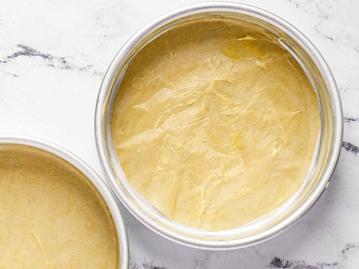 Two round pans lined with parchment and lightly greased, ready to bake a decadent gluten free dark chocolate cake, sit atop a white marble surface.