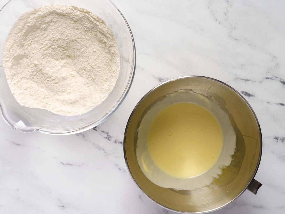 A glass bowl filled with flour sits next to a metal bowl containing a pale yellow liquid mixture-ingredients prepped for a gluten free dark chocolate cake-on a white marble surface.
