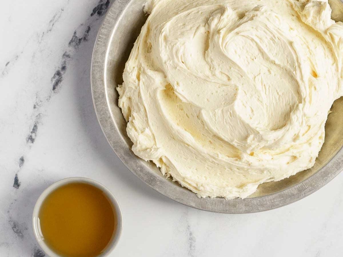 A metal bowl filled with creamy white frosting sits on a marble surface next to a small white bowl containing a yellowish liquid, perfect for topping your gluten free dark chocolate cake.