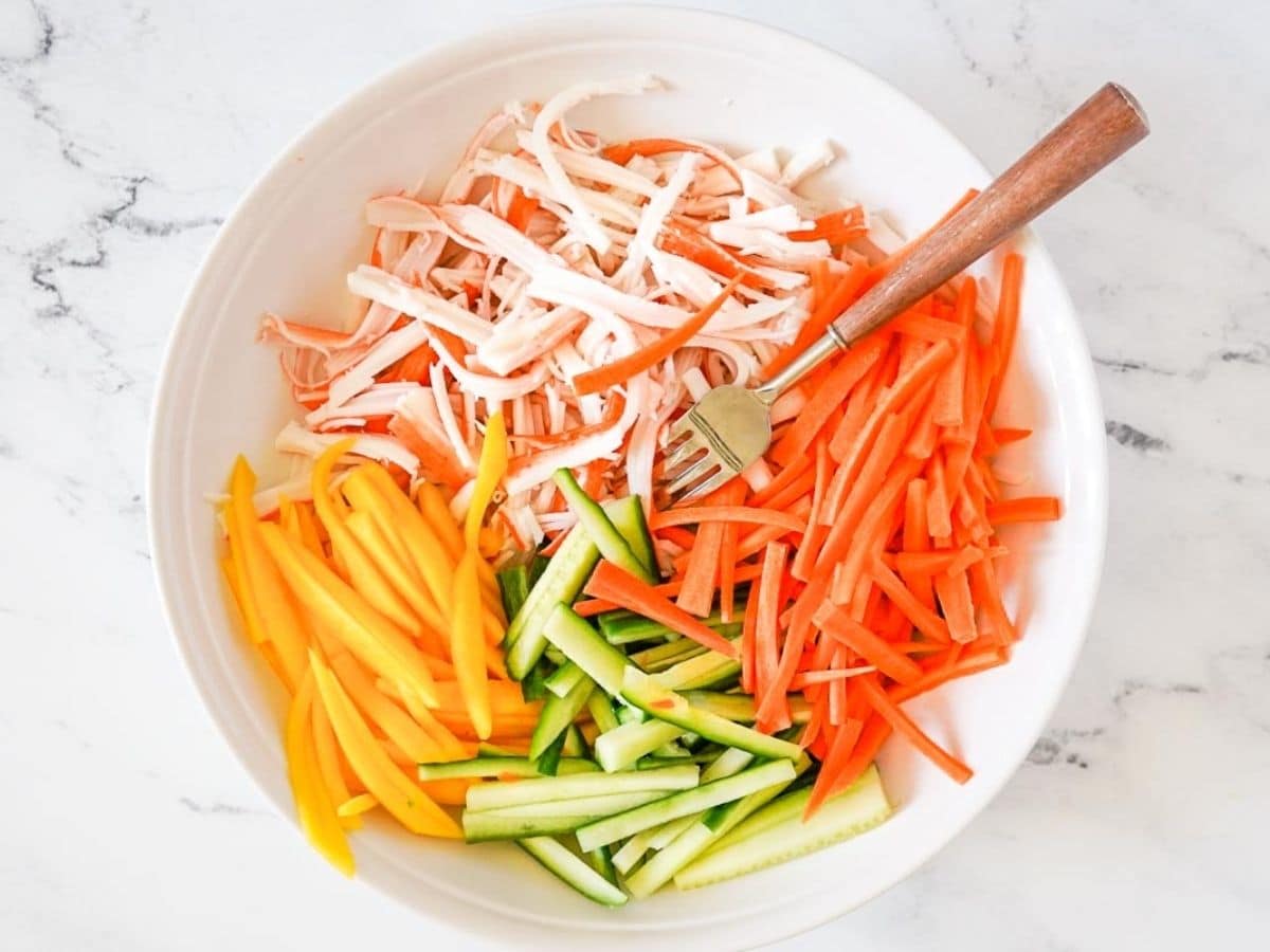 White plate with shredded imitation crab, carrot sticks, cucumber sticks, and yellow bell pepper sticks-perfect for making a spicy kani salad. Served with a fork on the side.