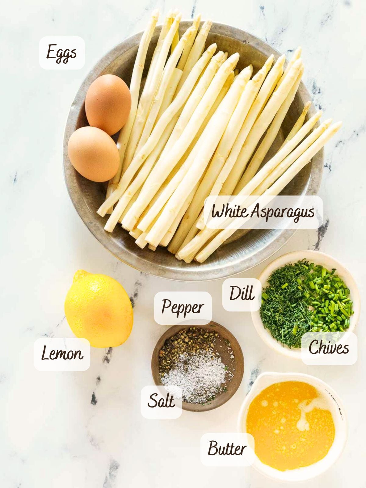 A bowl of eggs and white asparagus, perfect for your next white asparagus recipe, with labeled ingredients: lemon, salt, pepper, dill, chives, and melted butter on a marble surface.