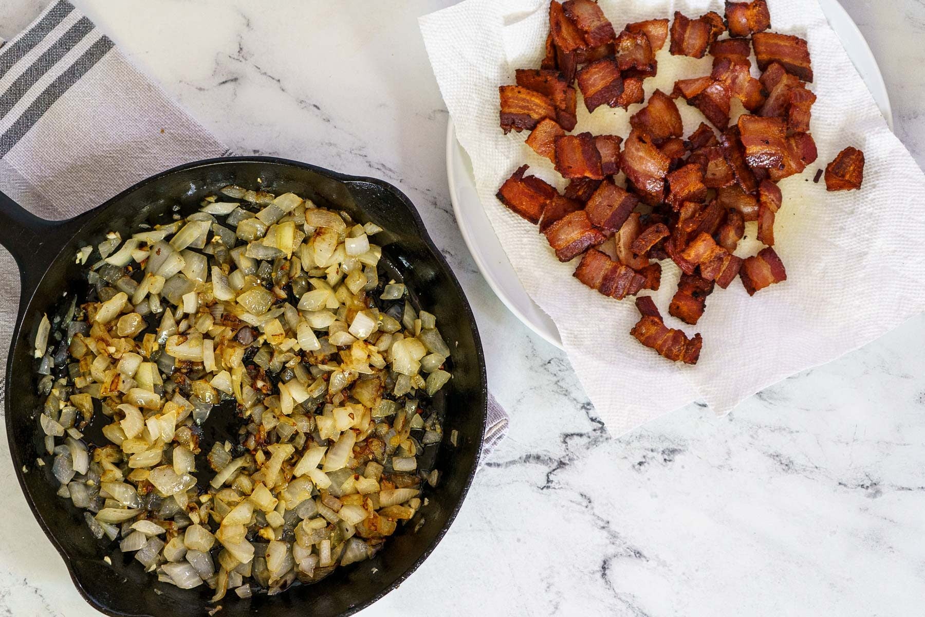 A cast iron skillet with sautéed onions sits beside a plate lined with paper towels holding cooked, chopped bacon-perfect ingredients for making molasses baked beans-on a marble countertop.