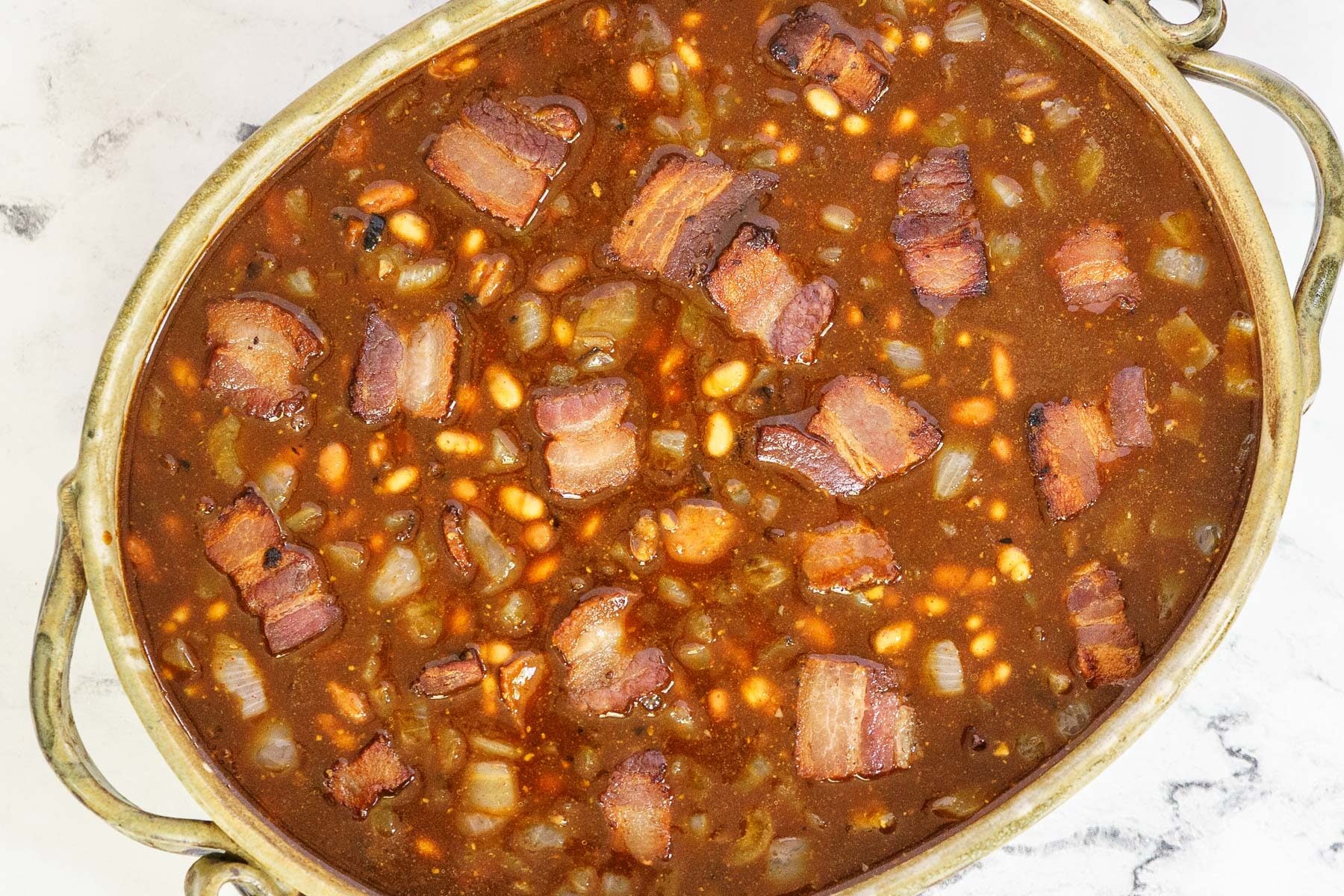 An oval dish filled with molasses baked beans in a rich sauce, topped with chunks of cooked bacon and chopped onions.