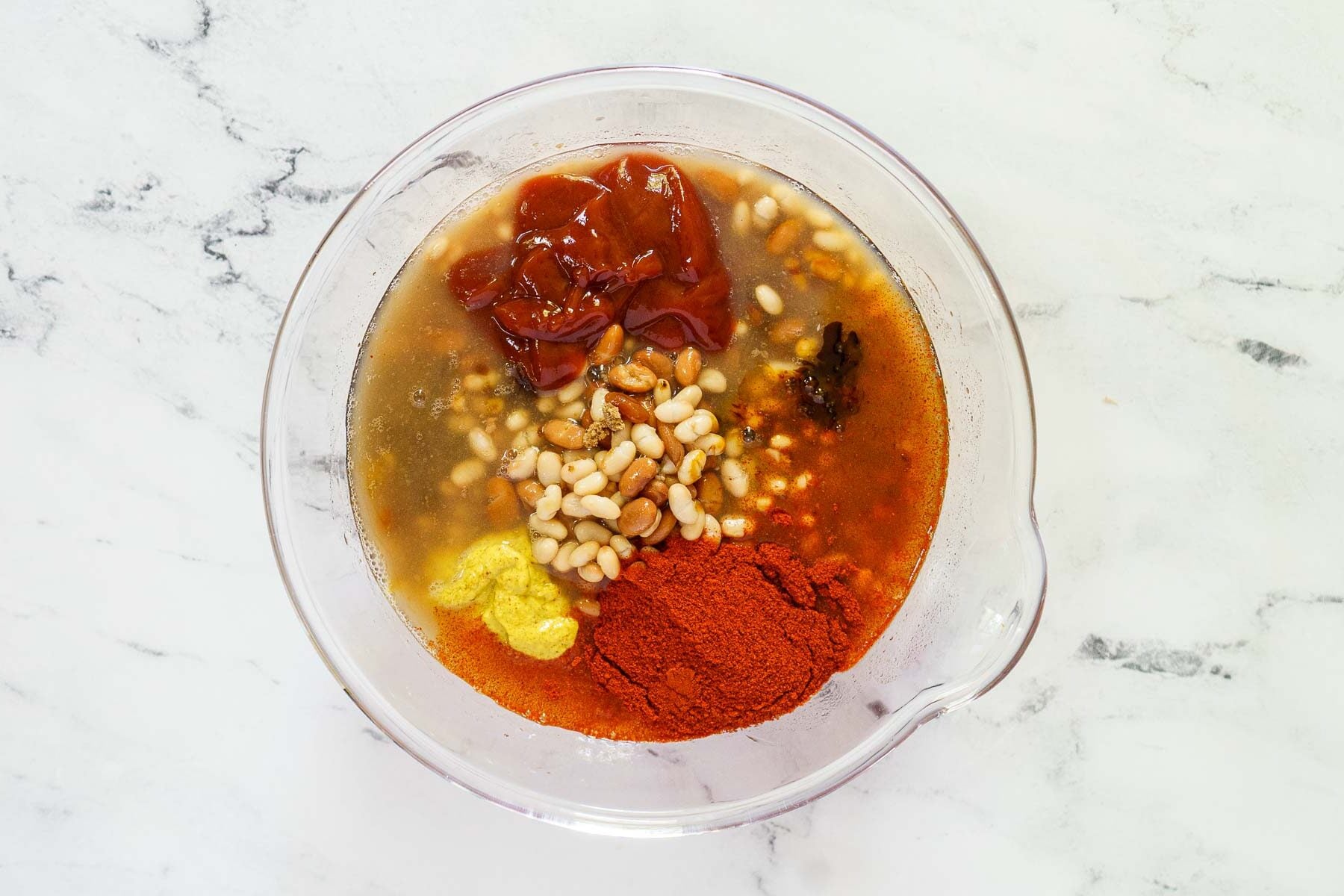 A glass mixing bowl containing ketchup, mustard, paprika, pine nuts, and other liquid ingredients-perfect for adding a rich twist to molasses baked beans-sits on a white marble surface.