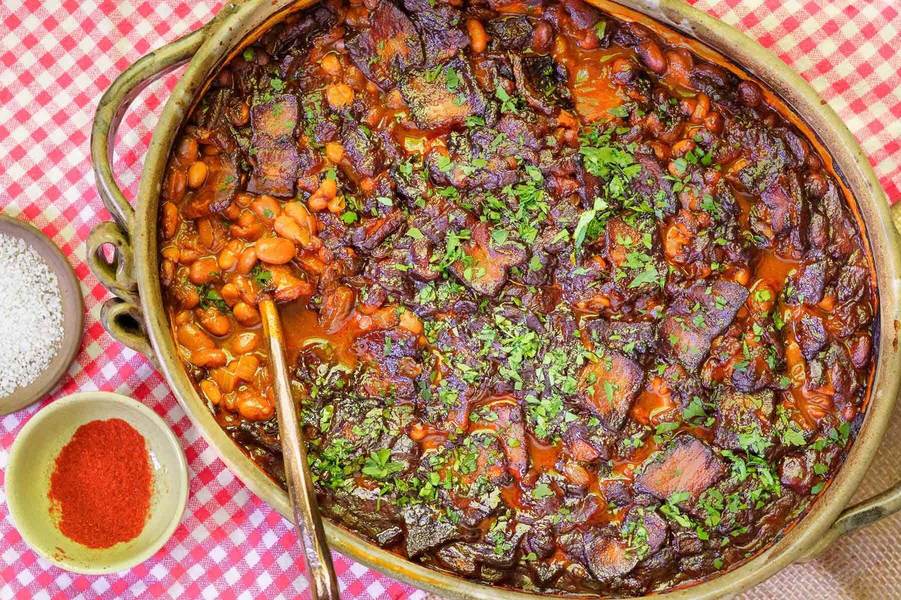 A large casserole dish of molasses baked beans in a rich tomato sauce, topped with chopped herbs, sits on a red and white checkered tablecloth next to bowls of salt and paprika.
