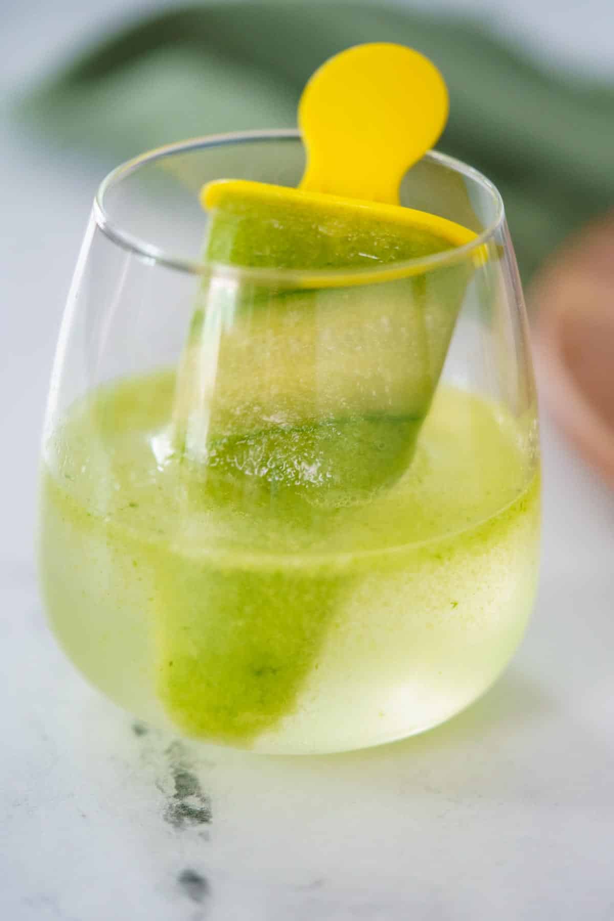 A green popsicle with a yellow stick is playfully nestled in a clear glass of cucumber gin and tonic, all set on a pristine white surface.