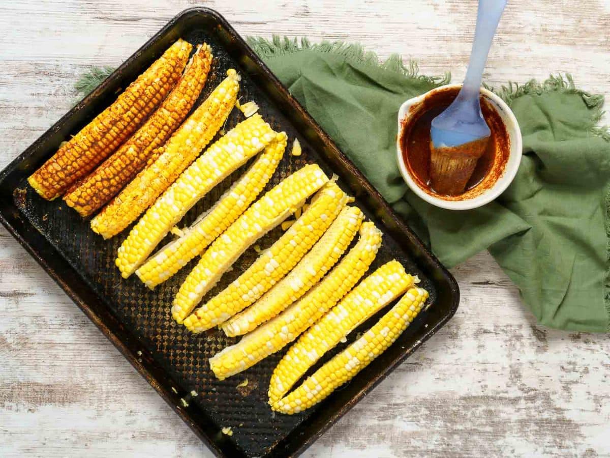 A baking tray with corn on the cob, some brushed with seasoning like a delicious corn ribs recipe, sits next to a bowl of sauce and a basting brush on a green cloth.