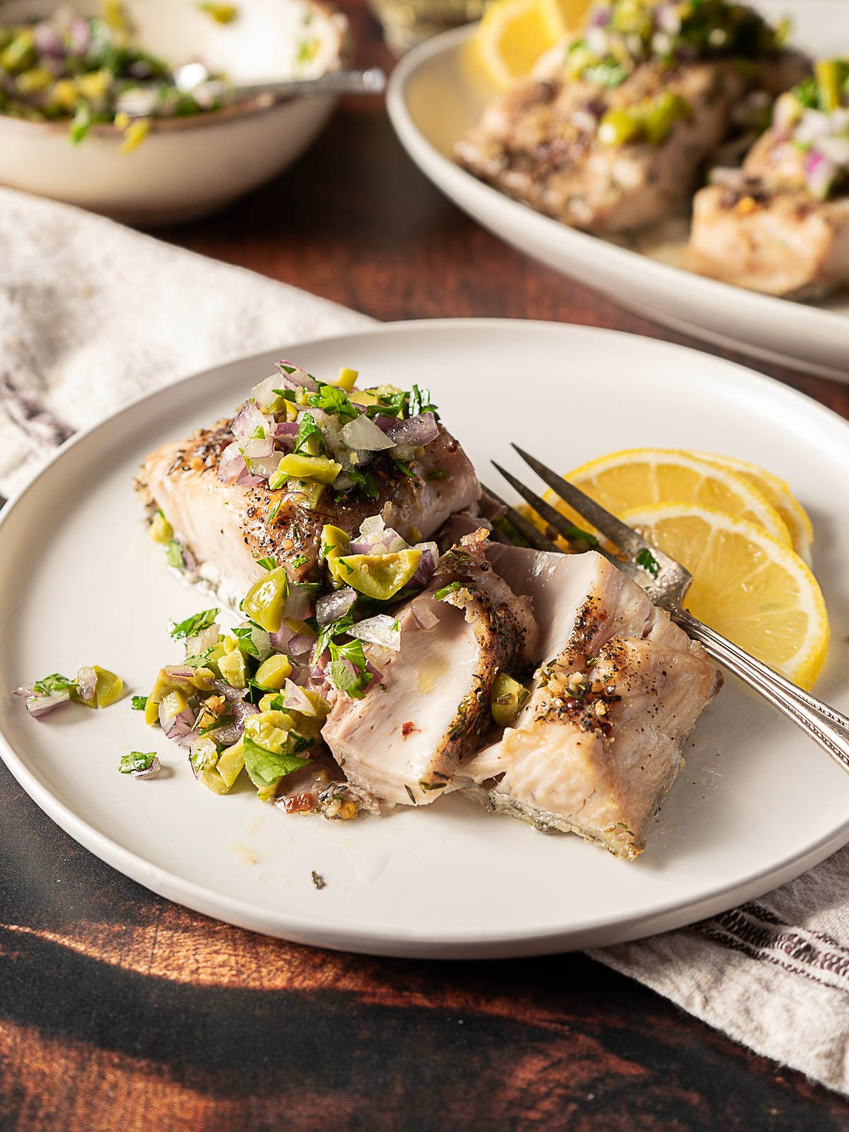Grilled fish fillet inspired by a baked mahi mahi recipe, topped with chopped green olives and herbs, served on a white plate with lemon slices and a fork.