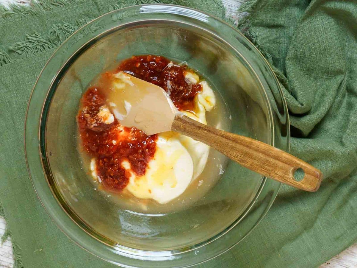 A glass bowl contains mayonnaise, chili sauce, honey, and minced garlic-ingredients perfect for a corn ribs recipe-with a wooden spatula resting inside, placed on a green cloth.