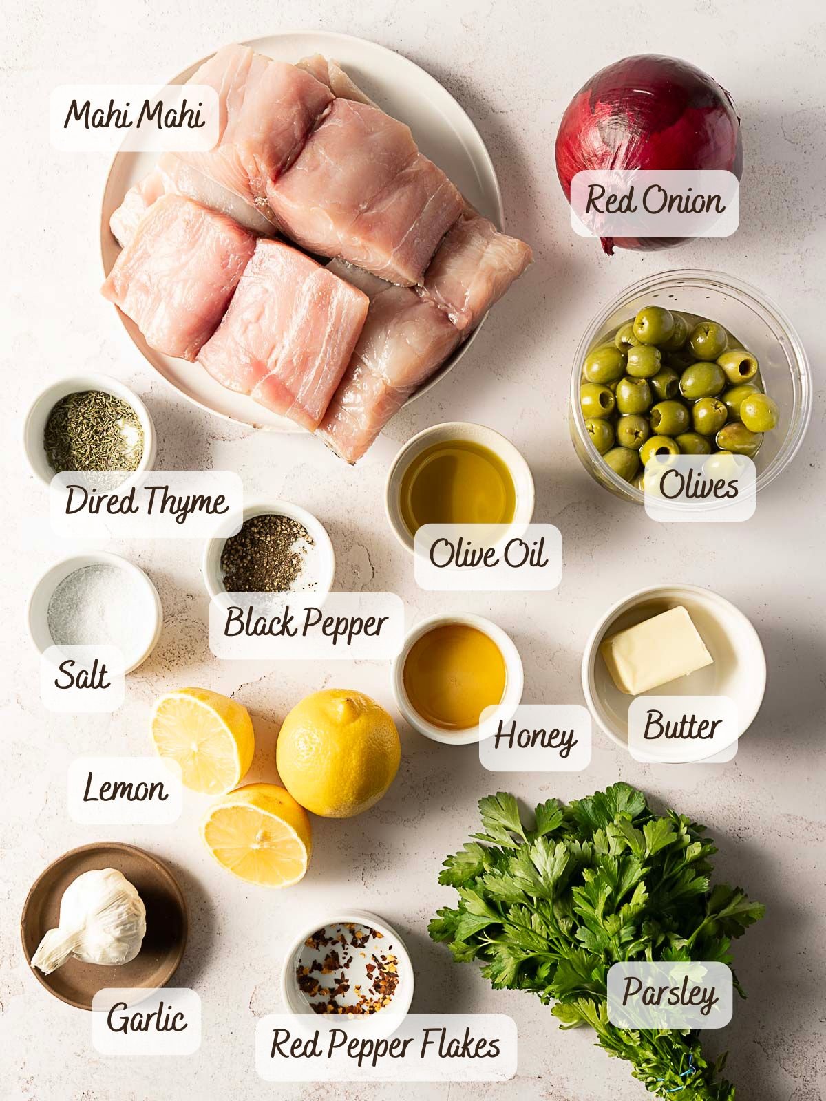 A flat lay of ingredients for a baked mahi mahi recipe, featuring mahi mahi, red onion, olives, dried thyme, olive oil, black pepper, salt, lemon, honey, butter, garlic, red pepper flakes, and parsley.