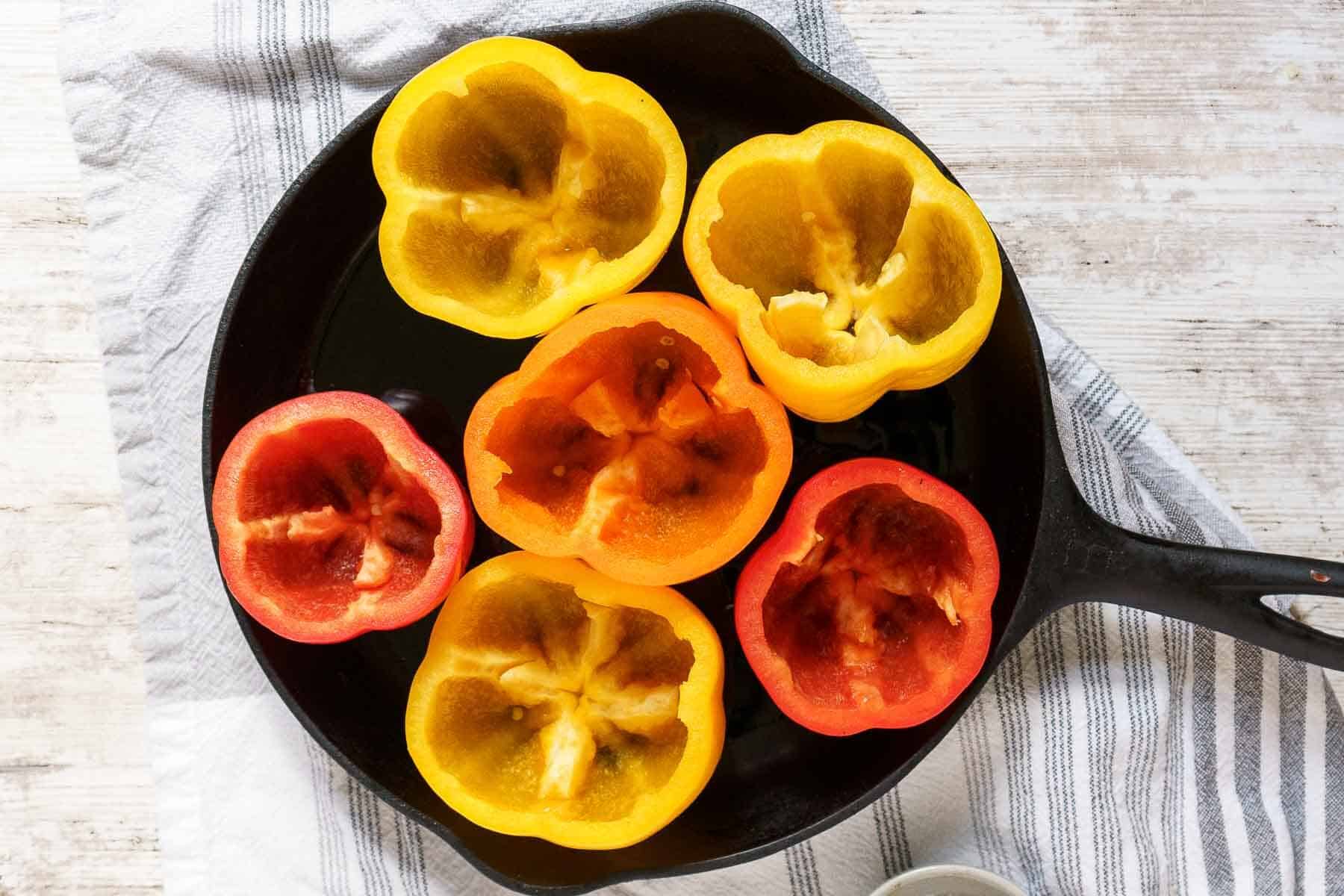 A cast iron skillet holds six ground chicken stuffed peppers-three yellow and three red halves-arranged cut side up on a striped cloth background.