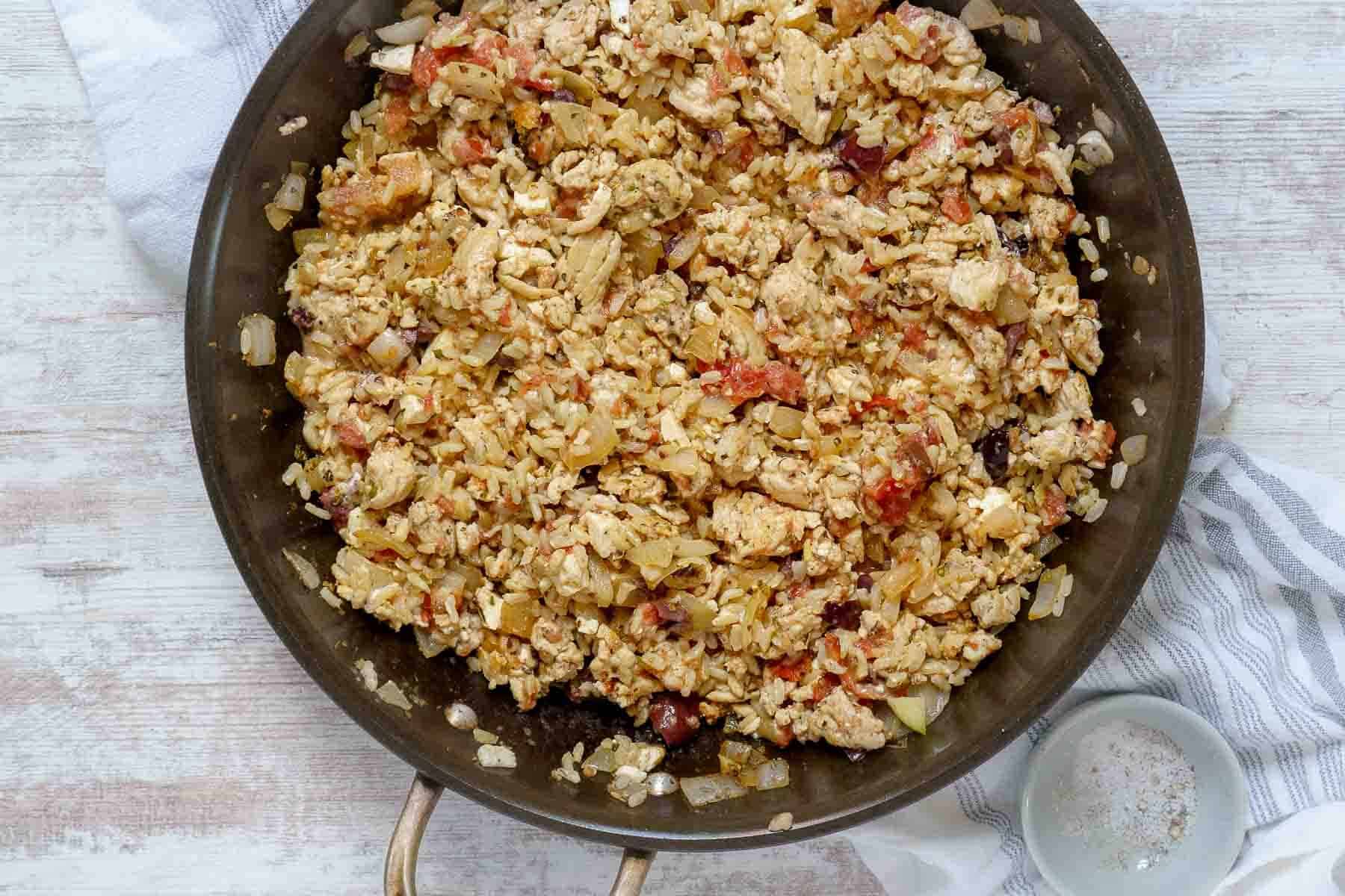 A skillet filled with scrambled eggs, diced tomatoes, onions, and a ground chicken stuffed peppers mixture sits on a white surface next to a small bowl of salt.