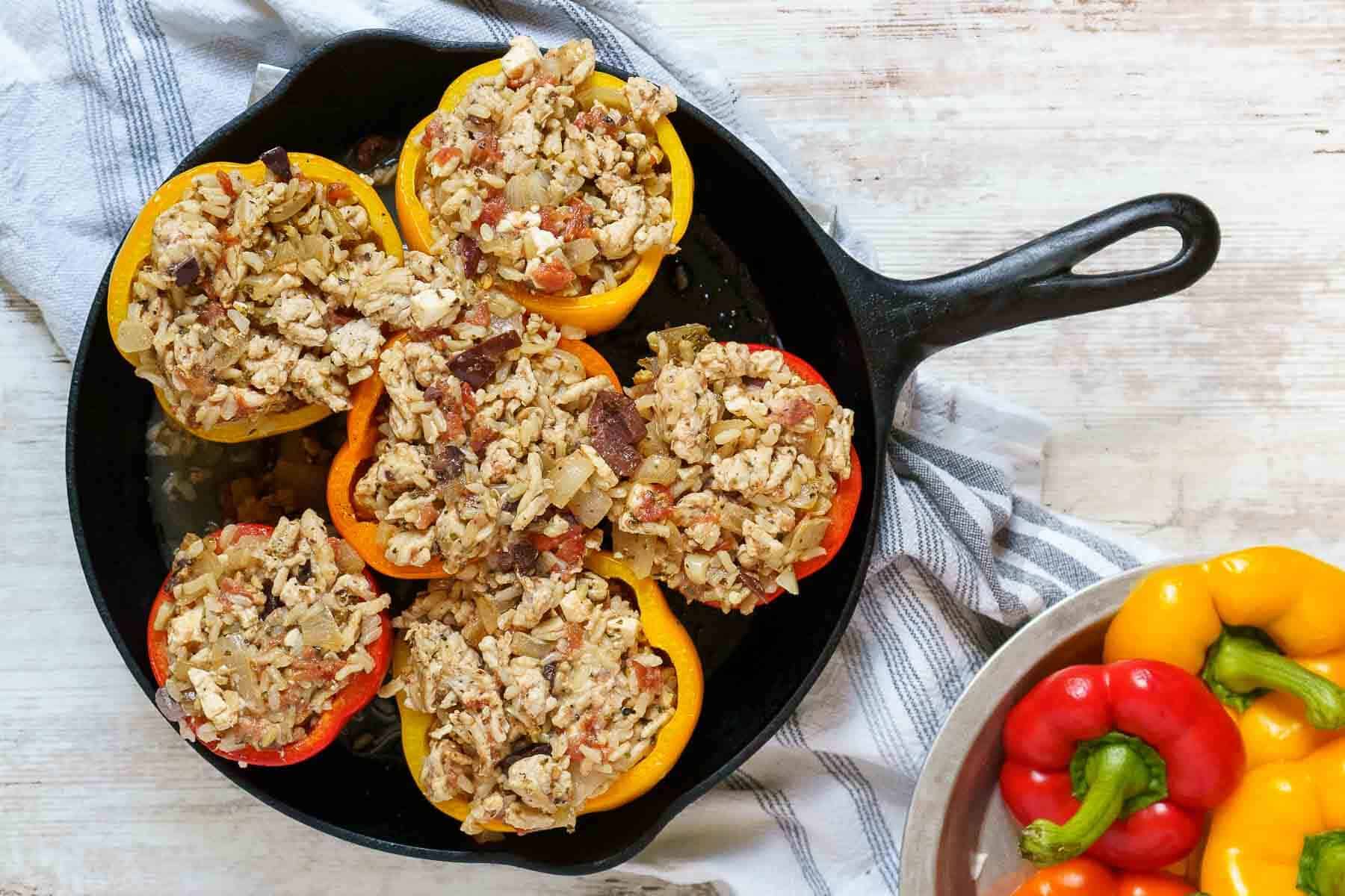 Ground chicken stuffed peppers filled with rice and vegetables are arranged in a cast iron skillet, next to a bowl holding whole red, yellow, and orange bell peppers.