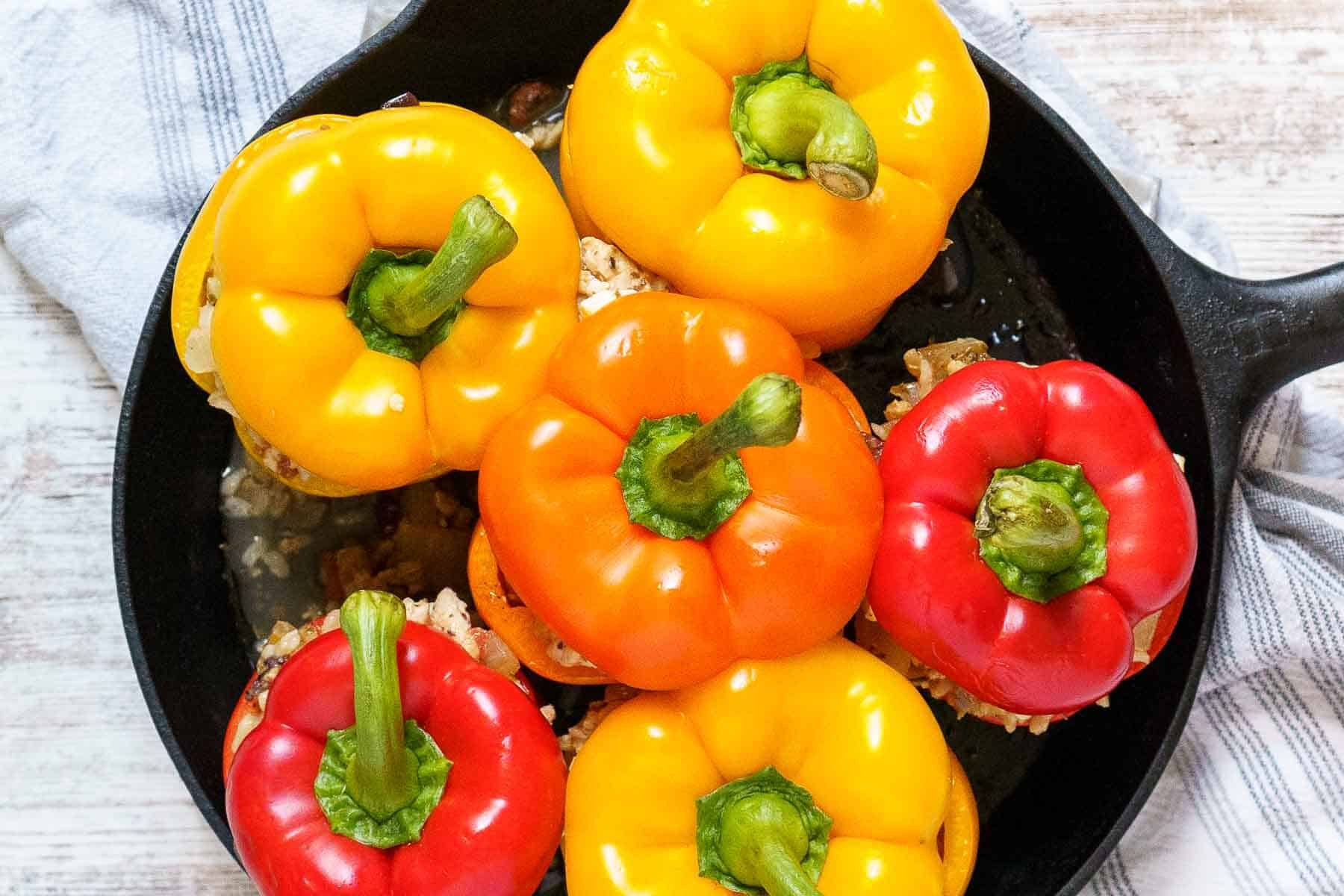 A cast iron skillet filled with vibrant ground chicken stuffed peppers in yellow, orange, and red hues, placed on a light-colored surface with a striped cloth nearby.