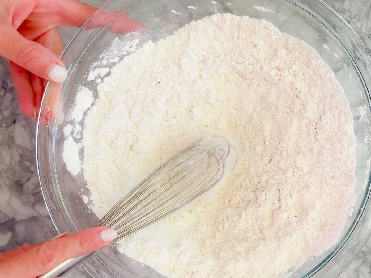 A person holds a clear glass bowl filled with flour mixture for a gluten-free lemon drizzle cake and uses a metal whisk to combine the dry ingredients. The bowl is resting on a gray marble surface.