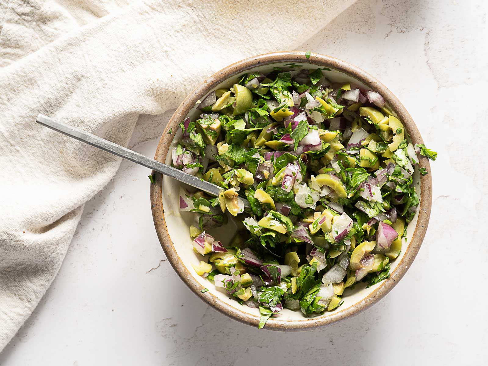 A bowl of chopped salad with green olives, red onion, and fresh herbs sits beside a metal spoon on a light surface-perfect as a refreshing side for your favorite baked mahi mahi recipe.