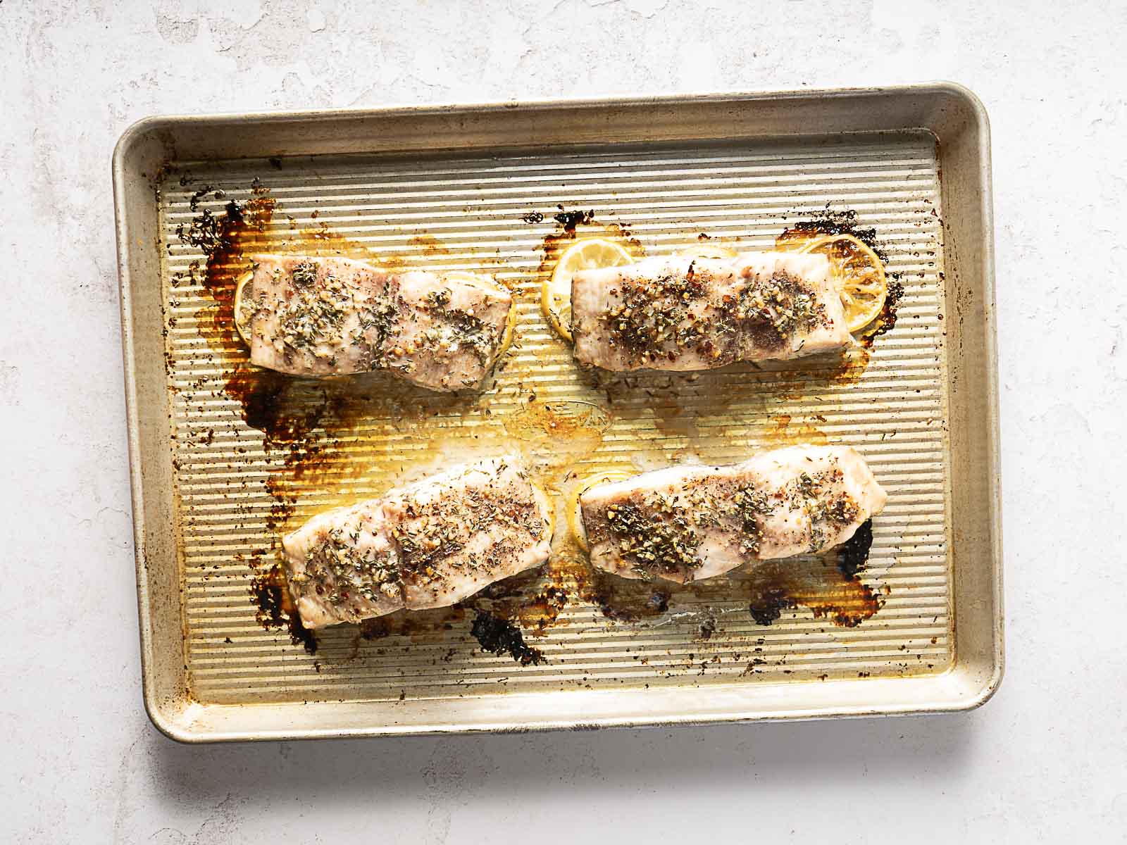 Four seasoned salmon fillets baked on a metal sheet pan, each fillet placed on top of a slice of lemon-an easy method that works just as well for a baked mahi mahi recipe.