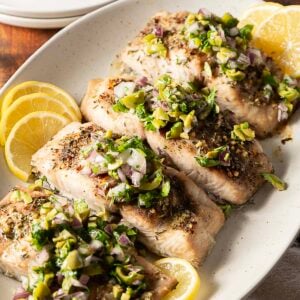 Four cooked salmon fillets topped with chopped olives, red onion, and herbs, served on a white plate with lemon slices-an easy dish to enjoy alongside your favorite baked mahi mahi recipe.