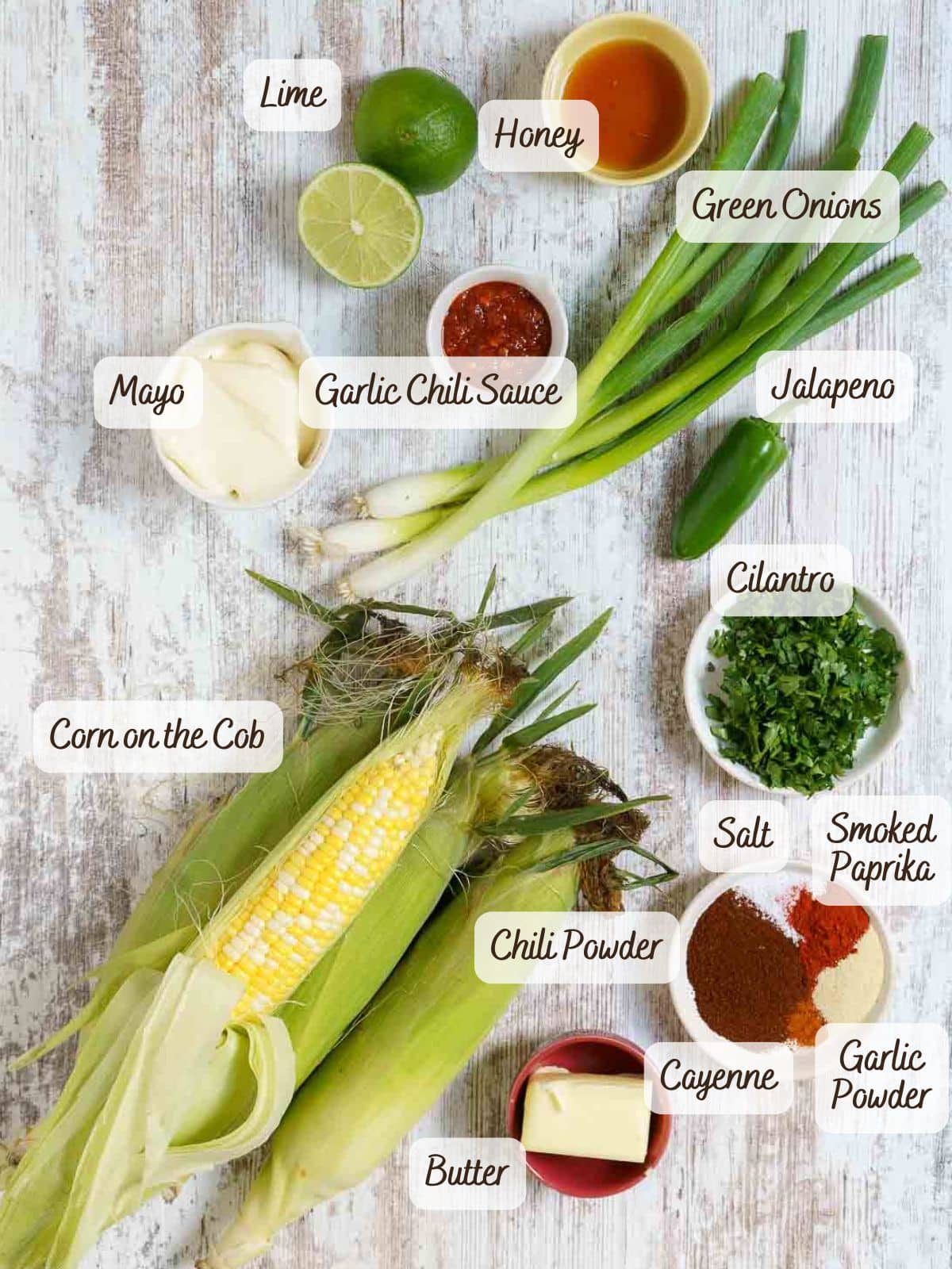 Ingredients for  corn ribs recipe, include corn on the cob, butter, mayo, lime, honey, green onions, jalapeno, cilantro, garlic chili sauce, spices, and salt-arranged on a white surface.