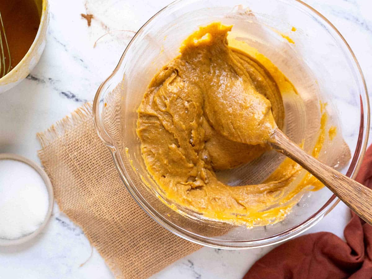 A glass bowl showcases a thick, orange batter for gluten-free pumpkin bread being stirred by a wooden spoon. The bowl rests on burlap, accompanied by a small container of white powder and a red cloth draped nearby.