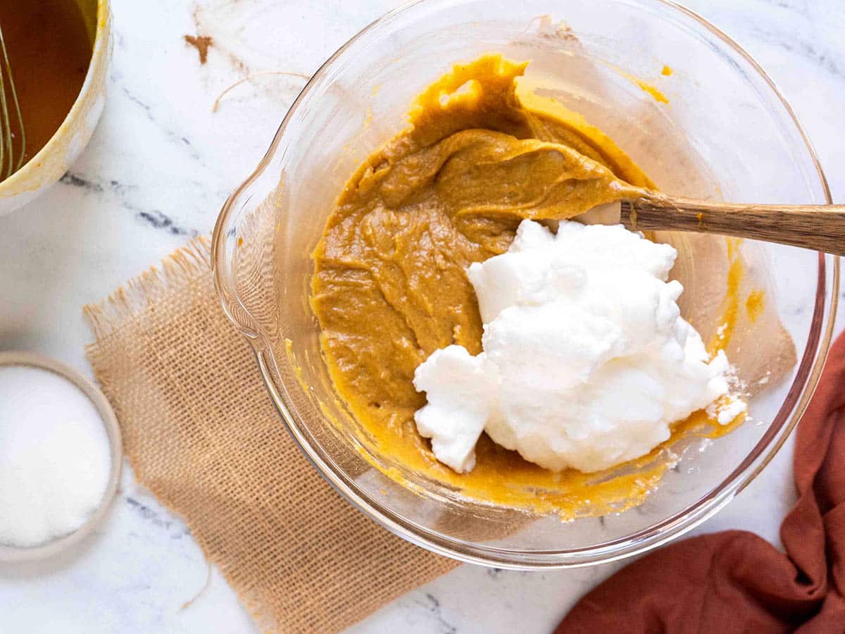 Glass bowl with gluten-free pumpkin bread batter and whipped egg whites being mixed on a white marble surface. A burlap cloth, small jar of sugar, and red cloth sit nearby, setting the perfect scene for baking.