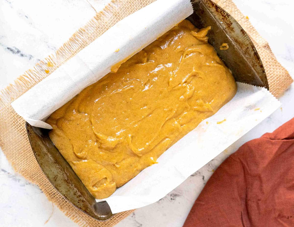 Gluten-free pumpkin bread batter in a parchment-lined loaf pan, ready for baking, placed on a burlap mat with a red cloth beside it.