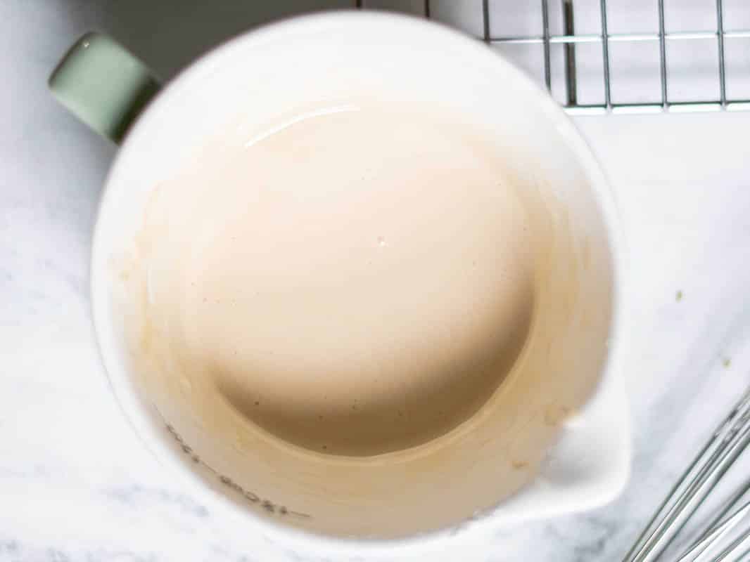 A measuring cup filled with a light beige batter for gluten-free pumpkin bread, sitting on a marble countertop next to a whisk and a cooling rack.