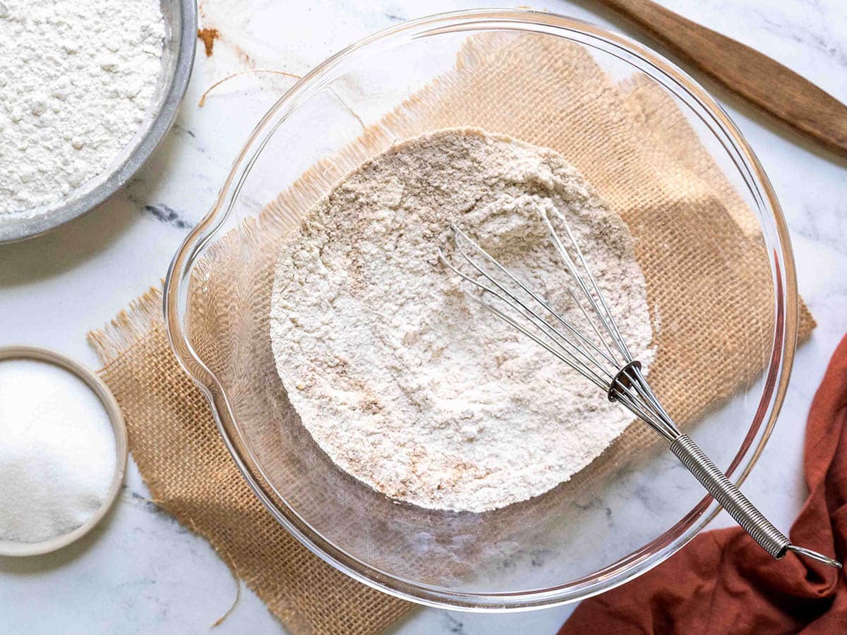 A glass bowl filled with flour and a whisk sits on a burlap cloth, ready for gluten-free pumpkin bread. Nearby, there are a dish of sugar, a wooden rolling pin, and a plate with additional flour.