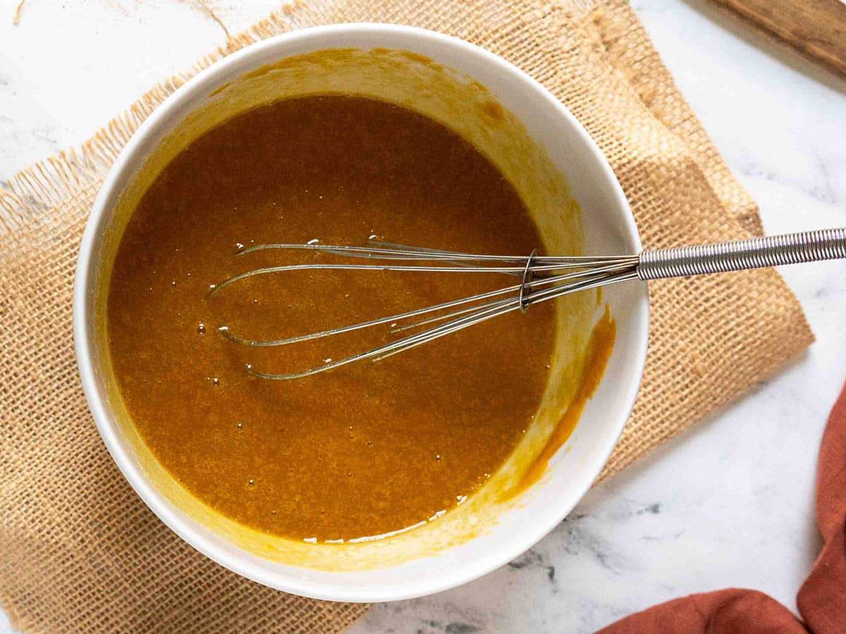 A white bowl containing a whisk and a thick, amber-colored mixture for gluten free pumpkin bread rests on a piece of burlap fabric on a marble surface.