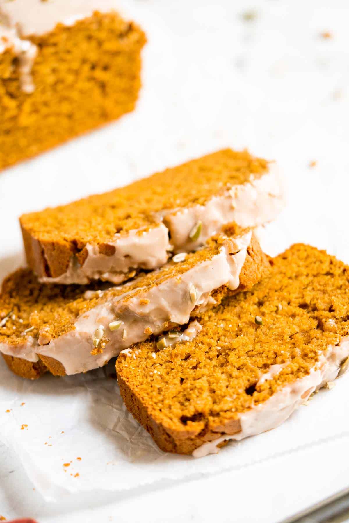 Three slices of glazed gluten-free pumpkin bread are stacked on a white surface with additional loaf pieces in the background.