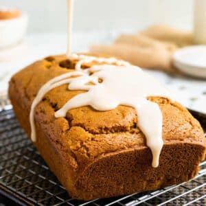 A glazed loaf of gluten-free pumpkin bread sits on a cooling rack, drizzled icing cascading down its sides.