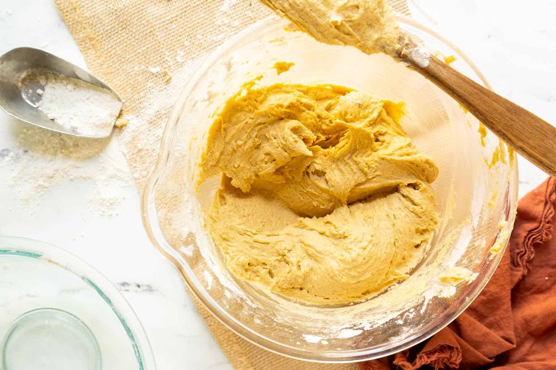 A glass mixing bowl filled with creamy gluten-free pumpkin cookie dough sits on a countertop next to a wooden spoon, a scoop of flour, and a brown cloth napkin.