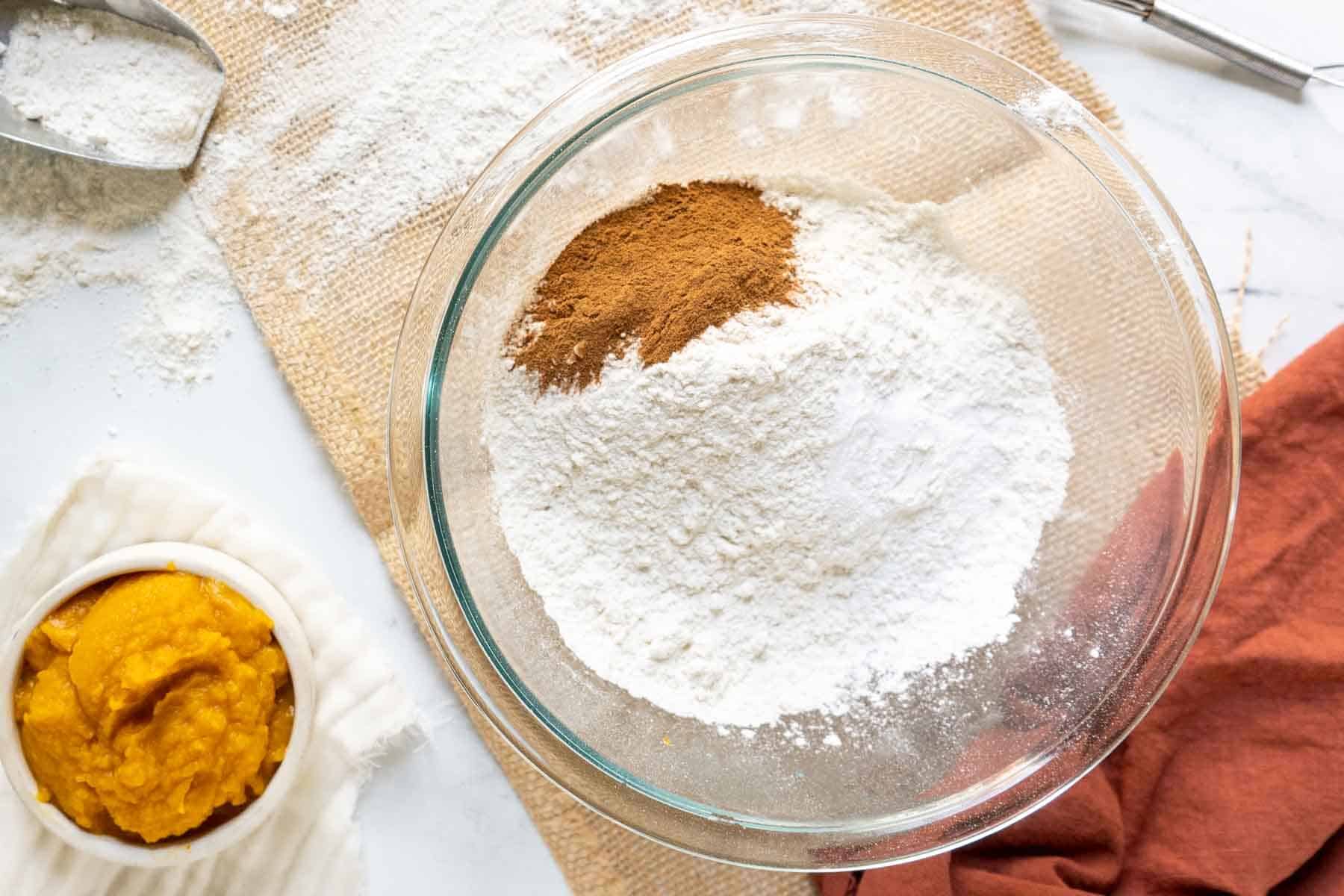 A glass bowl with flour and cinnamon on a table, next to a small dish of pumpkin puree-a perfect setup for making delicious gluten-free pumpkin cookies. A scoop with spilled flour completes the cozy baking scene.