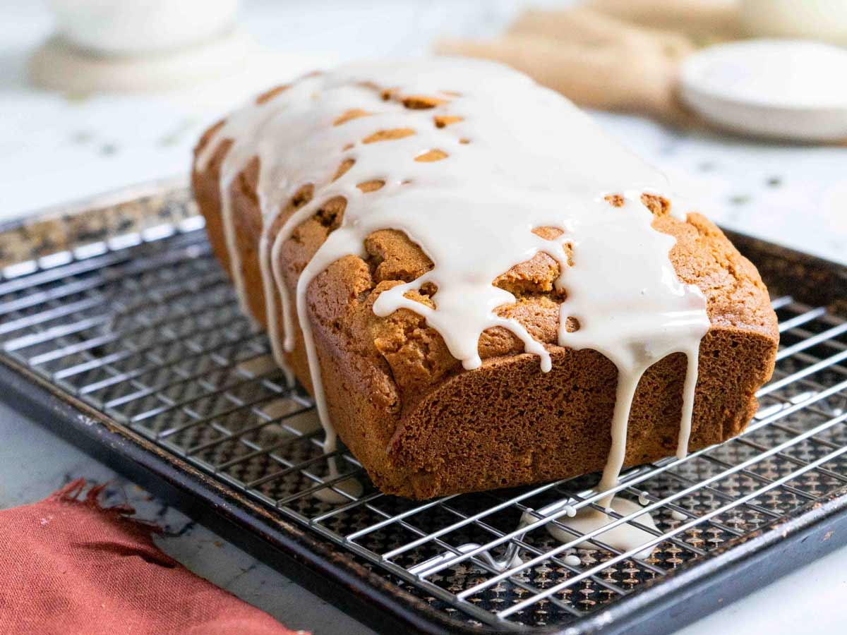 A loaf of gluten-free pumpkin bread sits on a cooling rack over a baking sheet, its fragrant glaze glistening under the light.