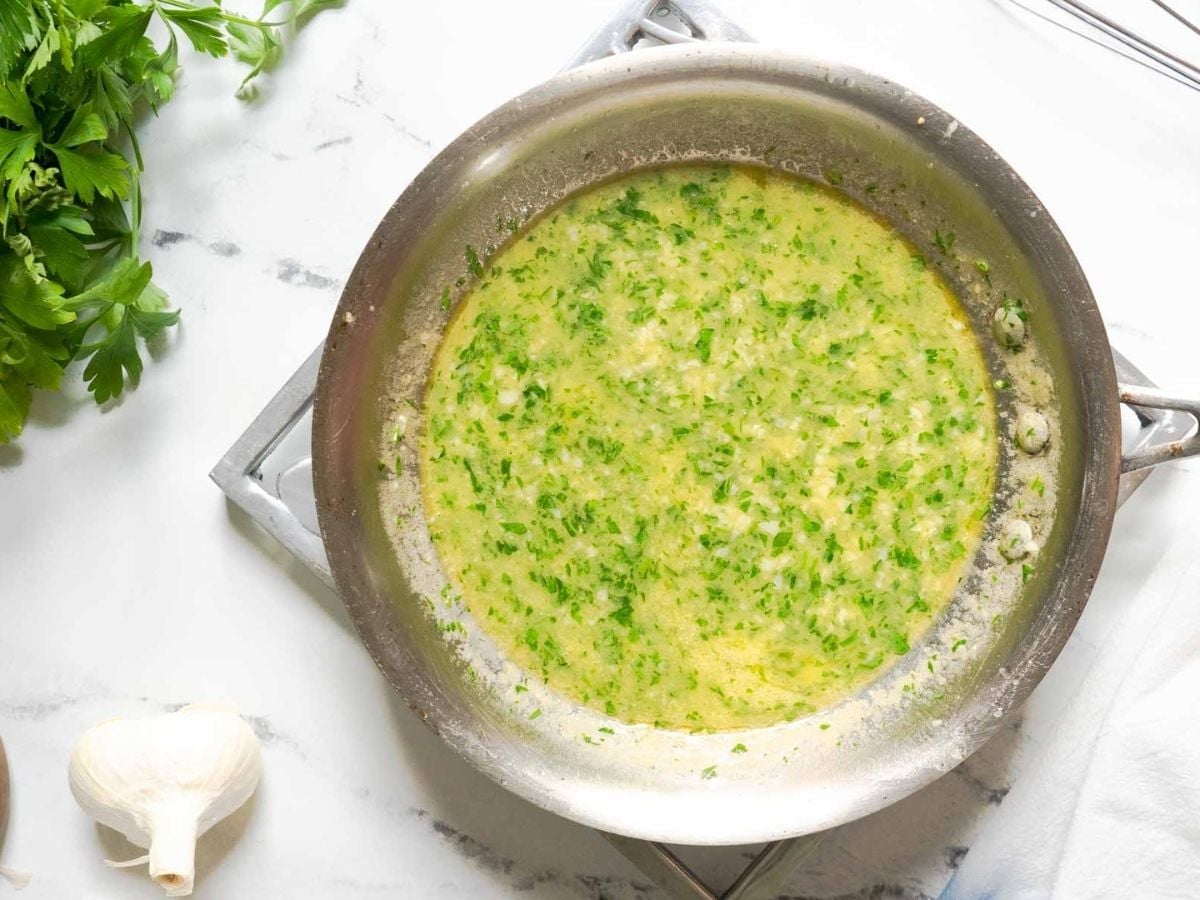 A pan filled with a creamy white wine garlic butter sauce sits on a stovetop, with fresh parsley and a garlic bulb nearby on a white marble surface.