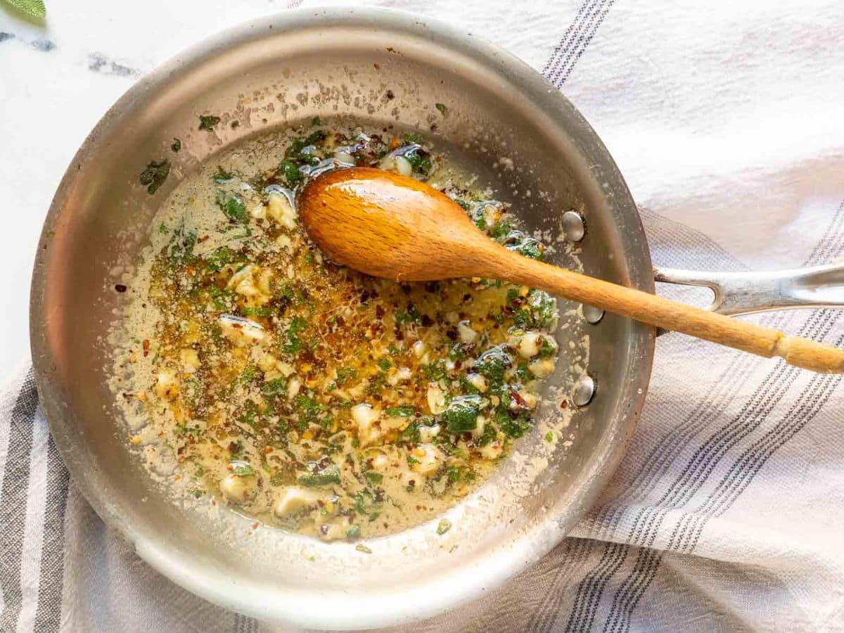 A stainless steel pan with sautéed garlic, herbs, and red pepper flakes in oil, stirred with a wooden spoon, sits on a white and striped kitchen towel-perfect for drizzling over Hasselback Butternut Squash.