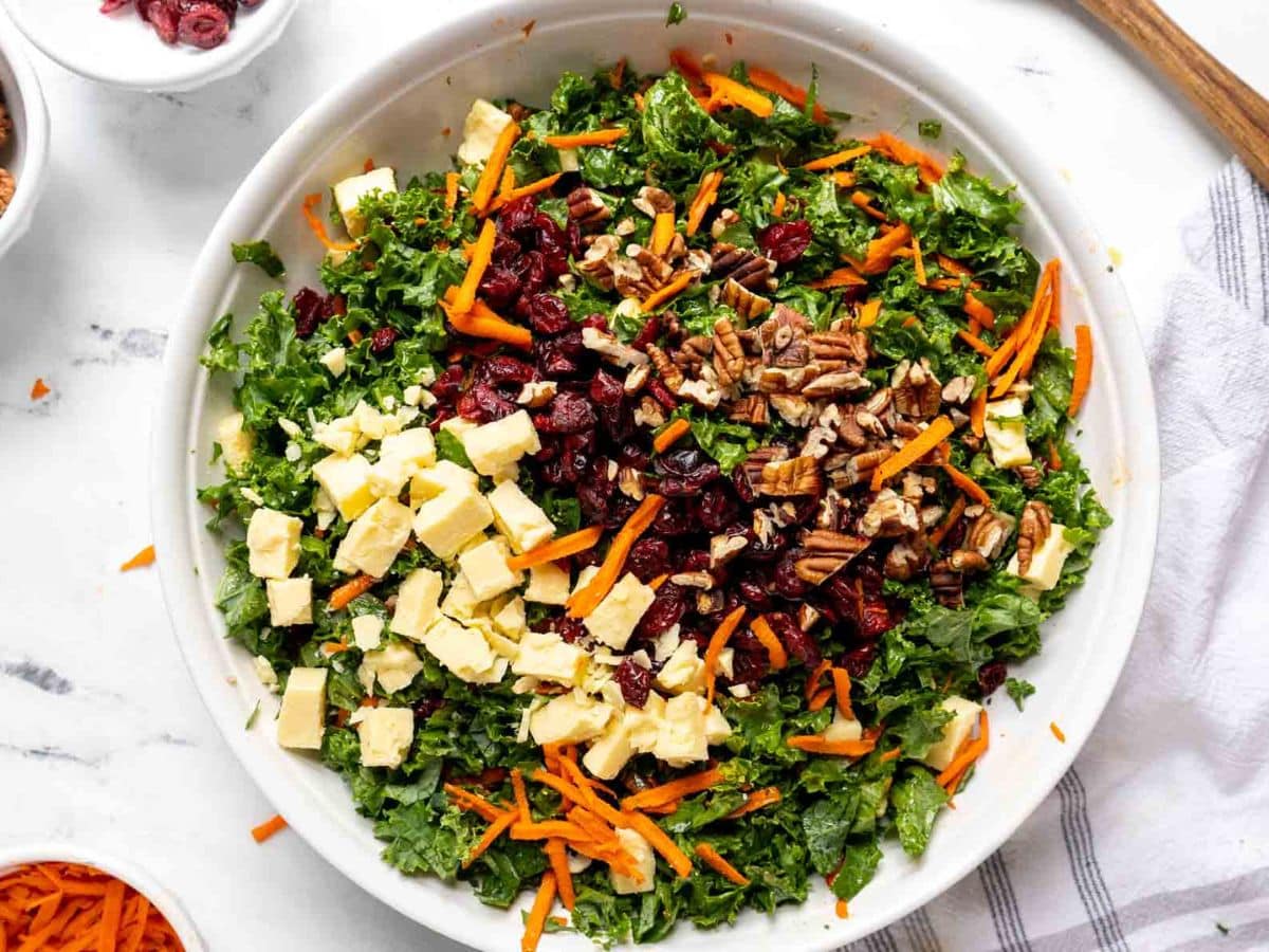 A bowl of Kale Salad with Cranberries features chopped kale, shredded carrots, dried cranberries, cubed cheese, and pecans, beautifully displayed on a white table.