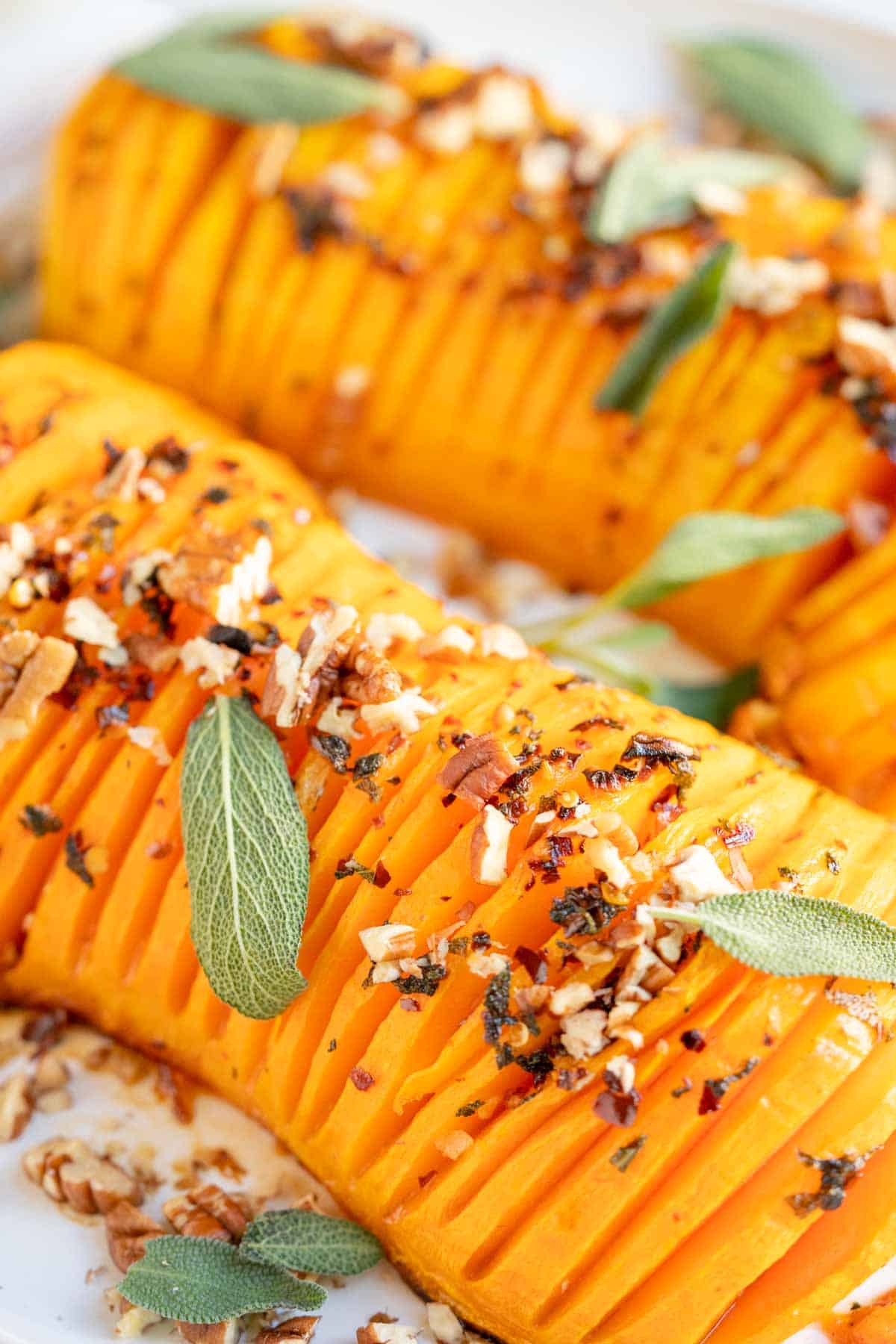 Hasselback squash on a white plate with sage leaves, pecans and chili flakes.