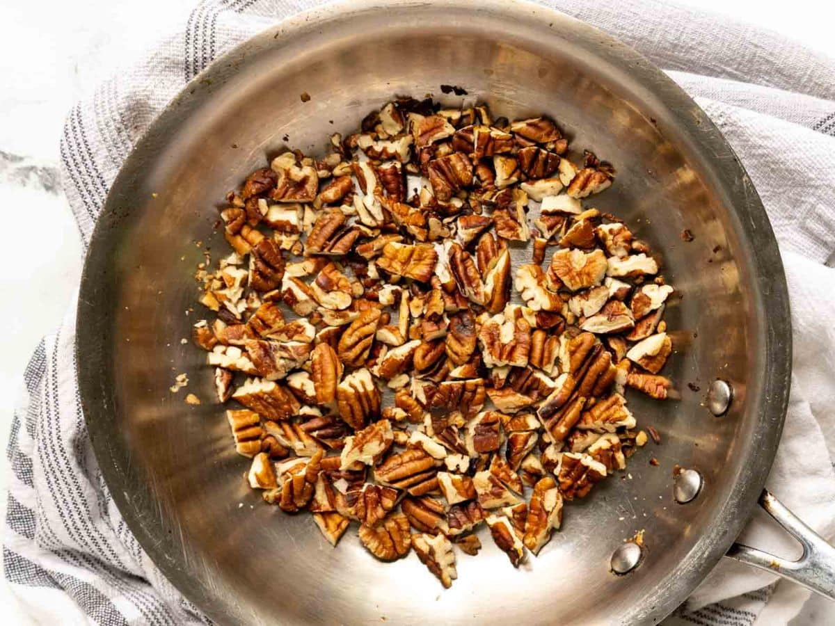 Chopped pecans being toasted in a stainless steel skillet on top of a gray and white striped cloth.