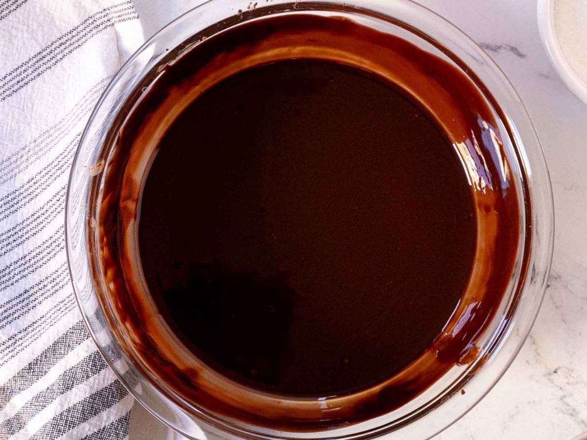 A glass bowl filled with melted chocolate, perfect for a chocolate souffle cake, sits on a white surface next to a white and gray striped kitchen towel. The chocolate is smooth and glossy, coating the sides of the bowl.