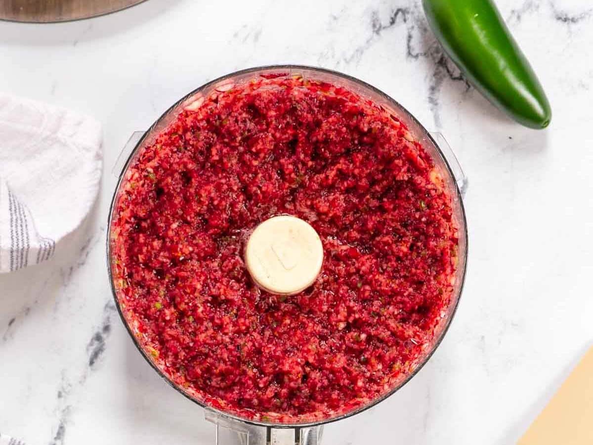 A food processor filled with finely chopped cranberry jalapeno dip sits on a white marble countertop next to a fresh green jalapeño and a striped towel.