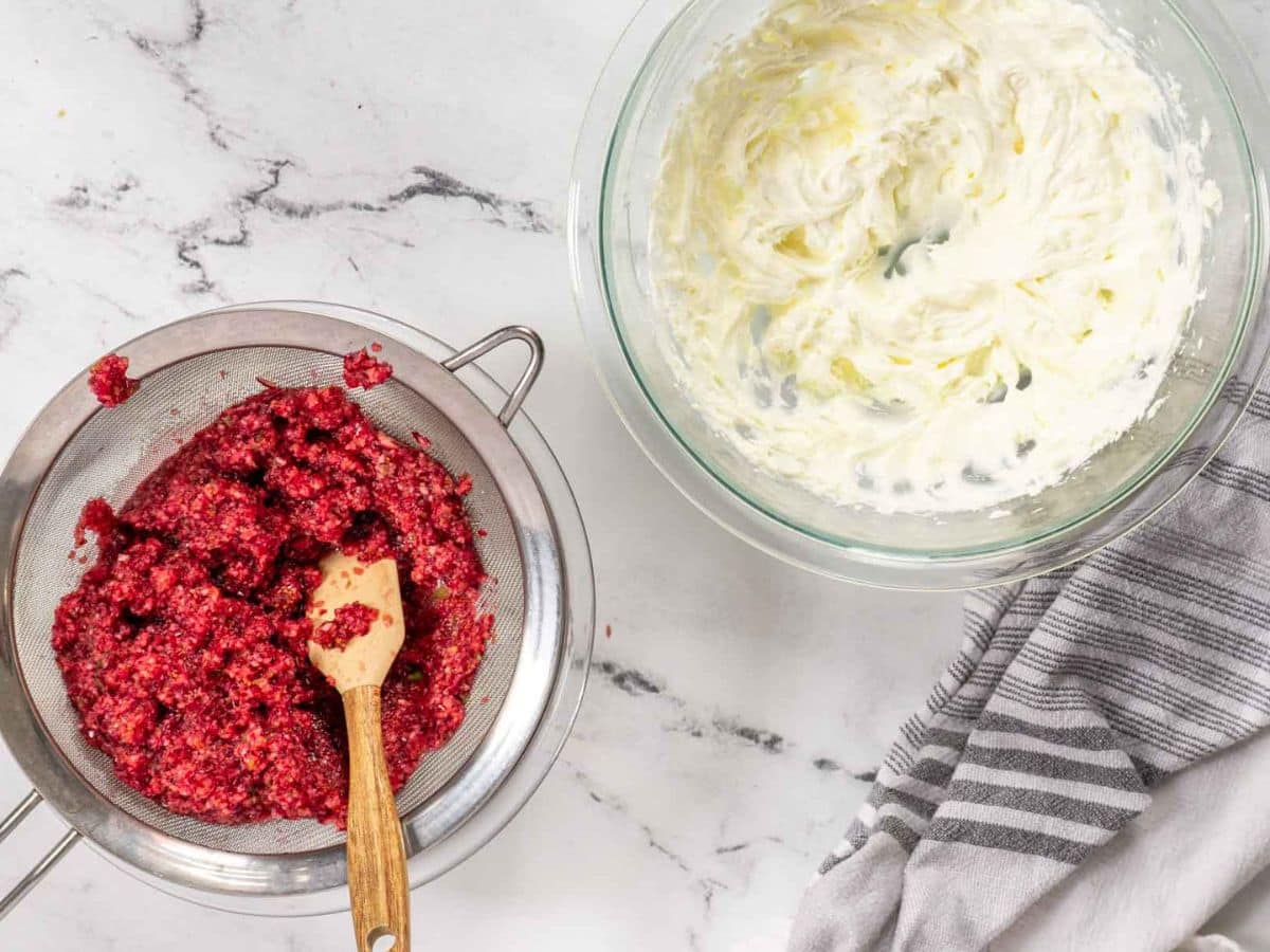 A strainer with mashed cranberries and a wooden spoon sits next to a bowl of whipped cream, all on a marble countertop with a striped towel.
