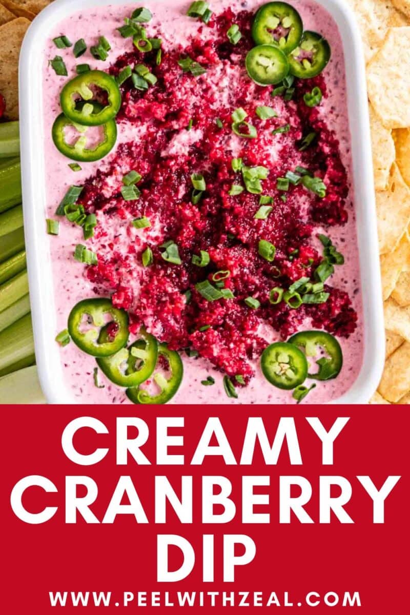 Rectangular dish of creamy cranberry jalapeno dip topped with chopped cranberries, sliced jalapeños, and green onions, surrounded by celery and chips.