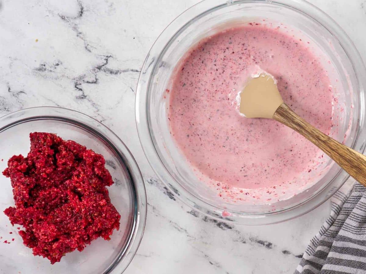 Two glass bowls on a marble surface: one with pink cranberry jalapeno dip batter and a spatula, the other with a mound of minced cranberries draining. A striped cloth is partially visible.