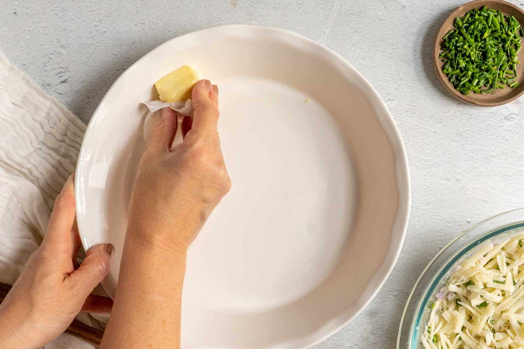 Hand greasing a white pie dish with butter, preparing for a delicious zucchini crustless quiche. Chopped herbs and grated ingredients sit nearby on a light surface, ready to join the culinary masterpiece.