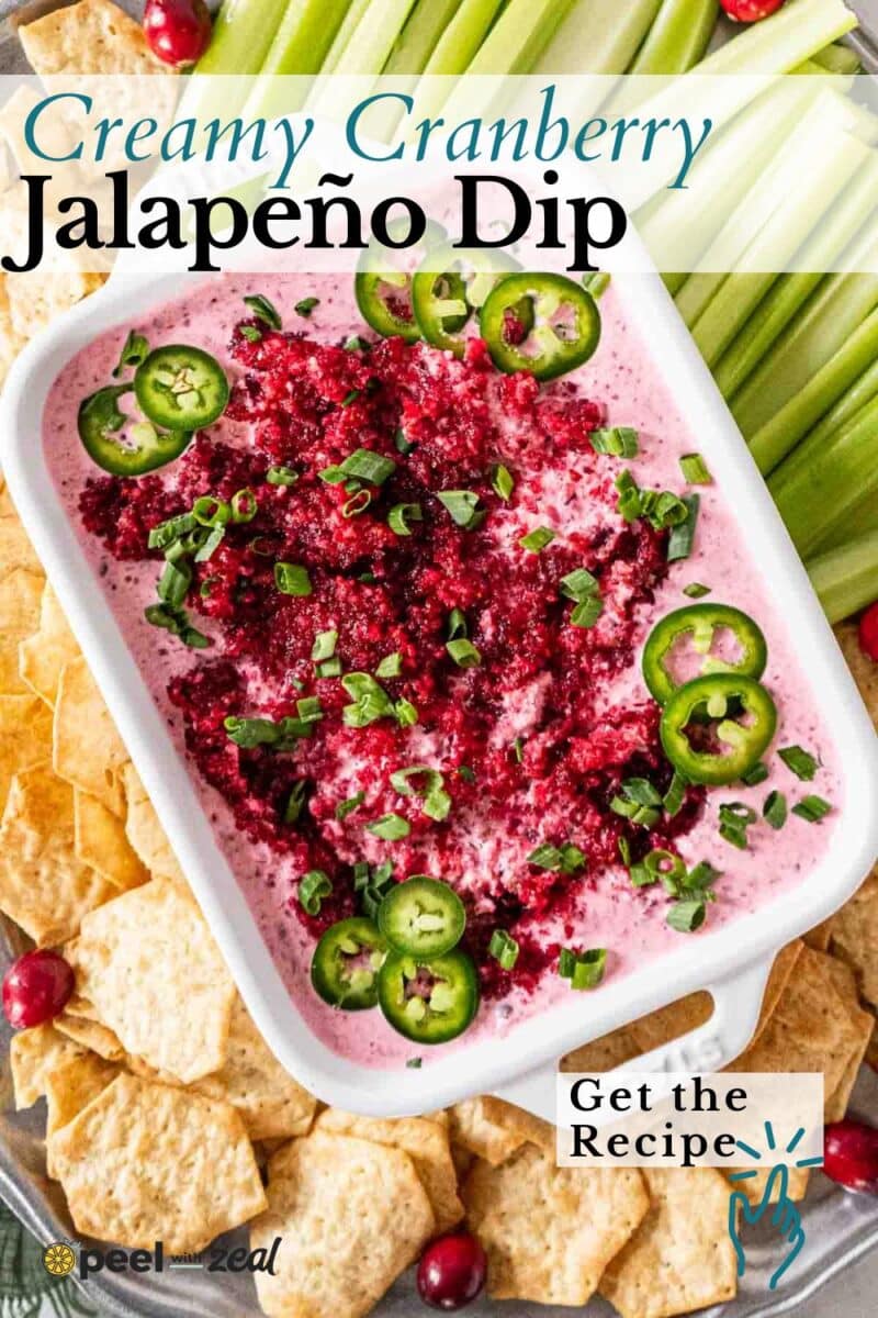 A rectangular dish of creamy cranberry jalapeno dip, garnished with sliced jalapeños and green onions, is surrounded by celery sticks and crackers.