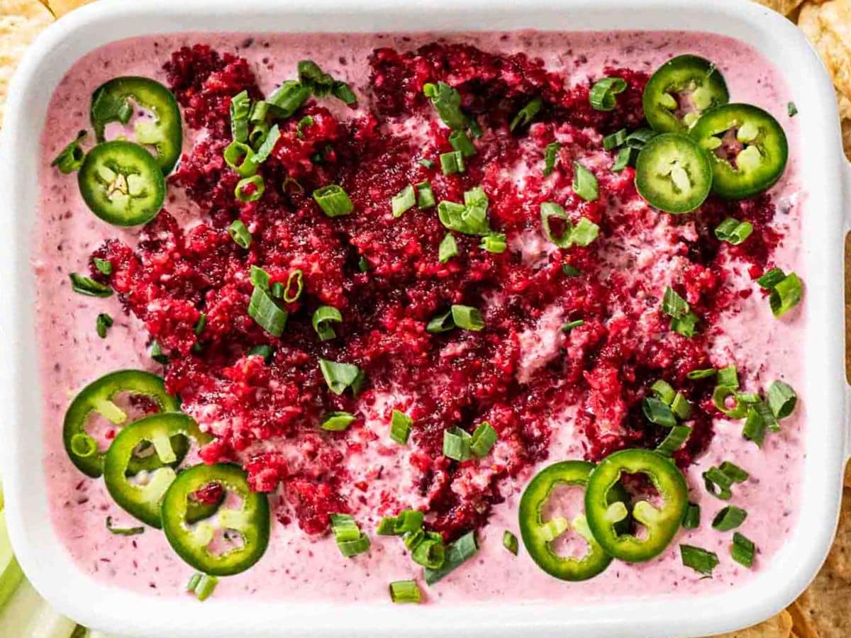 A rectangular dish filled with creamy cranberry jalapeno dip, topped with chopped green onions, jalapeño slices, and a finely chopped red garnish.